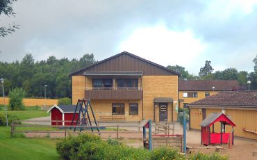 Bild på junibackens förskola med lekplatsen i förgrunden. huset är av gult tegel och brunt trä och är i två våningar