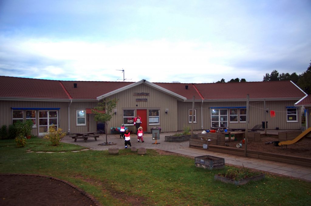 Bild på Ängsgårdens förskola som är ett grått enplanshus med röd entrédörr.