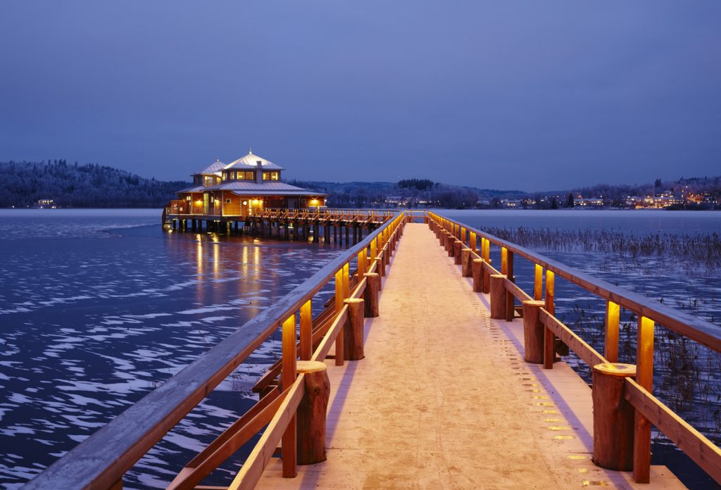 Bild på Kallbadhuset i Ulricehamn. Bilden är tagen kvällstid där lamporna lyser upp träbron ut till kallabdhuset som står på pålar i vattnet.