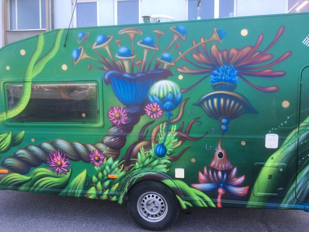 Bild på en husvagn målad i grön bakrund med konstnärliga figurer och växter.