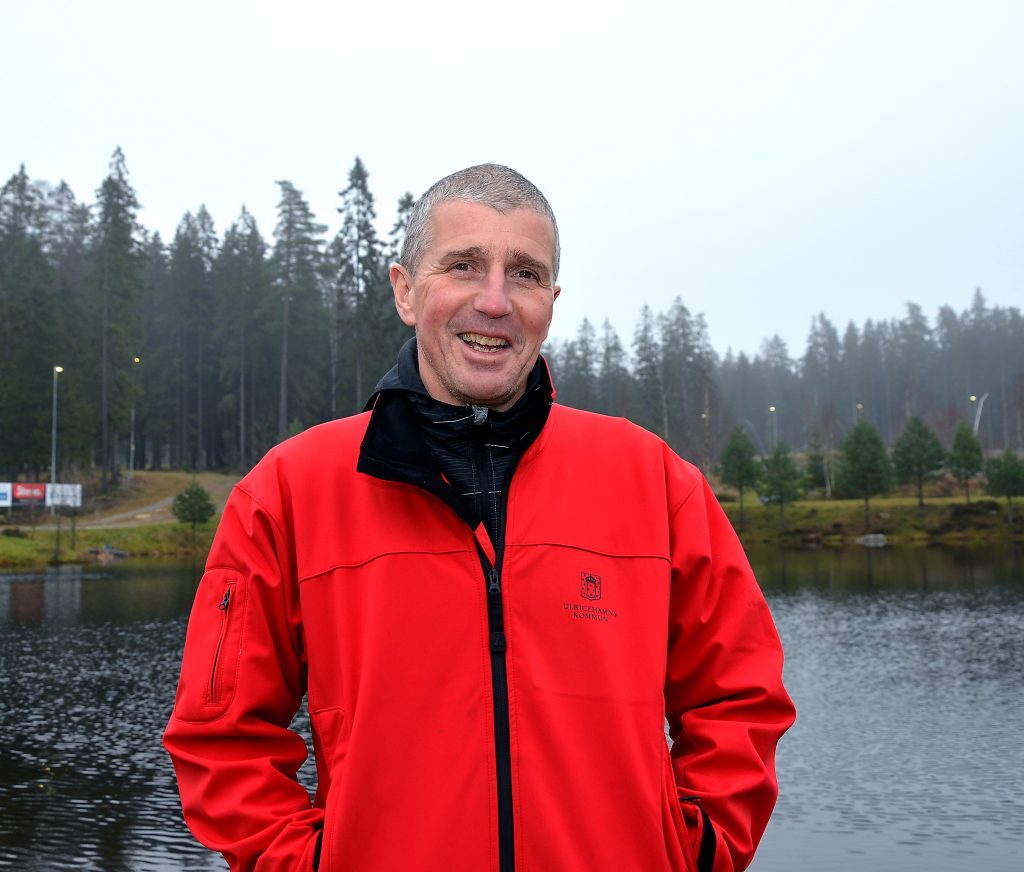 Bild på en man i 50-årsåldern som står vid en damm i röd jacka. Han ler.