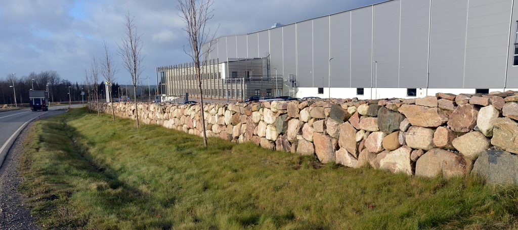 Bild på stenmur längs en väg och bakom den en industribyggnad utan fönster.
