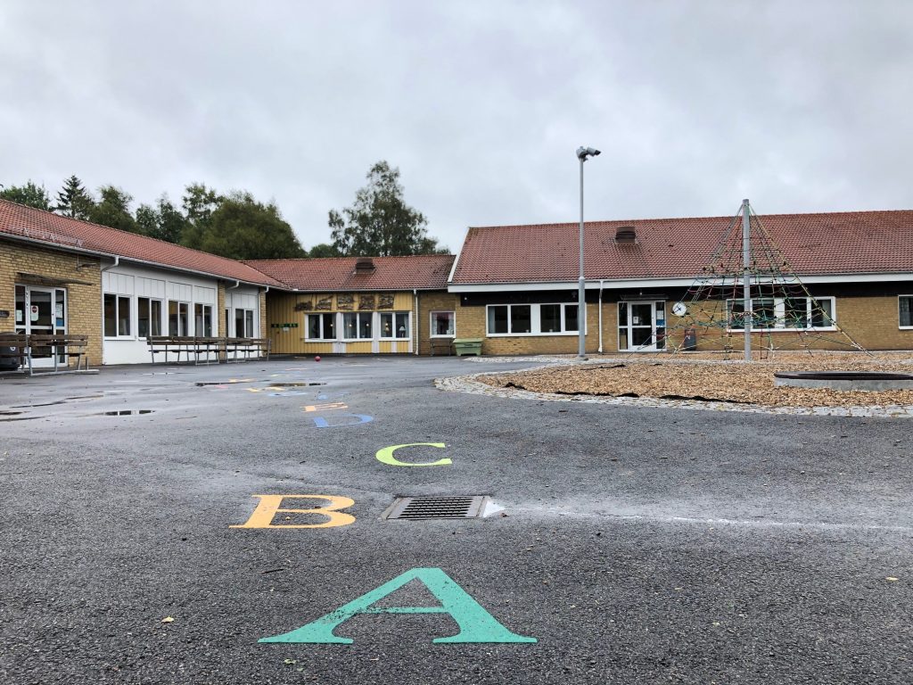 Bild på Hössna skola i gult tegel med asfaltsbeklädda skolgården i förgrunden