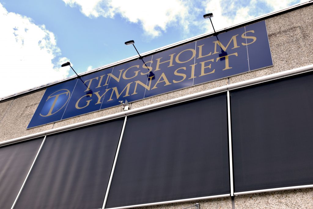 En blå skylt på en grå byggnad. På skylten står det Tingsholmsgymnasiet.