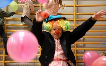 En clown i regnbågsfärgad peruk åker rutschkana nerför en bänk lutad mot en ribbstol i en gympasal. I luften flyger ballonger och i bakgrunden klättrar barn på ribbstolarna.