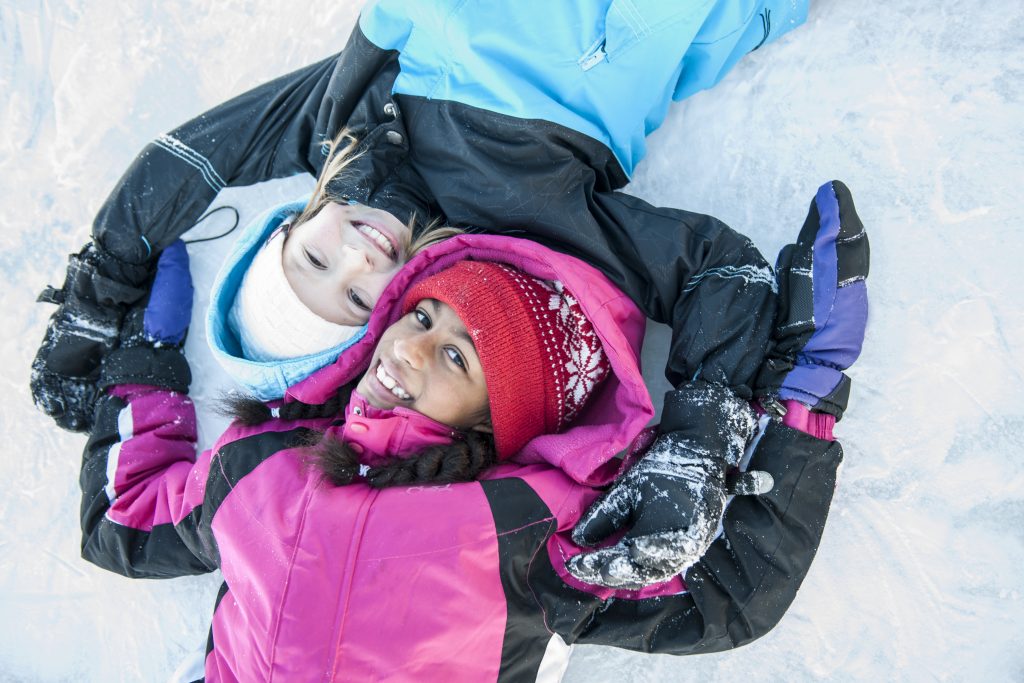 Två tjejer i vinteroveraller och mössor ligger huvud mot huvud i snön, båda är glada.