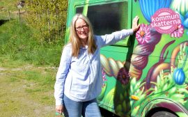 Kulturstateg Vicki Klaiber står lutad mot kommunens läshusvagn. Som är målad i grönt och med blommor och växter i olika färger klara färger. Vicki har långt blont hår, glasögon och är iklädd ljusblå skjorta.