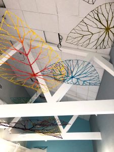 Bild på hängande konstverk i taket på en skolbyggnad. Konstverket består av textil och ståltråd och föreställer löv i flera olika färger.