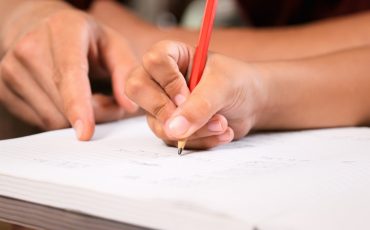 En lärare hjälper en elev. Eleven håller i en penna och skriver på ett papper. Läraren pekar på det eleven skriver.