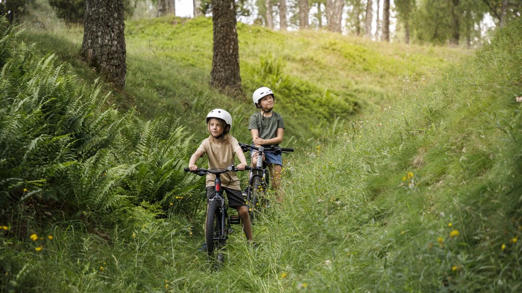 Två barn, en pojke och en flicka står med sina cyklar på en av hålvägarna i Timmele. Båda har vita hjälmar och tittar framåt, uppåt. Det är sommar och de är omgivna av grönska