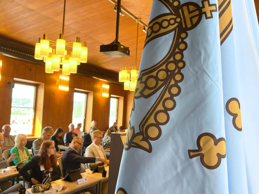 I förgrunden syns Ulricehamns kommuns flagga. I bakgrunden sitter människor i kommunfullmäktigesalen.