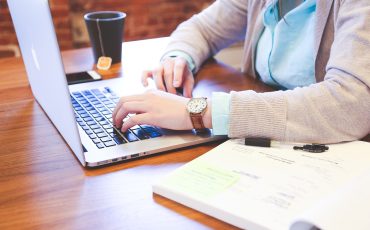 En person sitter vid ett bord och skriver på en laptop. Jämte datorn står en kopp te och en bok