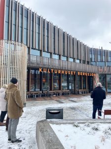 Bild på kulturhuset utifrån. Fasaden är träklädd, stora orange bokstäver med orden Kulturhuset Bergsjön. På marken ligger ett tunt lager av pudersnö.
