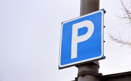 En närbild på en blå skylt med ett stort P på. Skylten sitter på en stolpe vid en parkering.